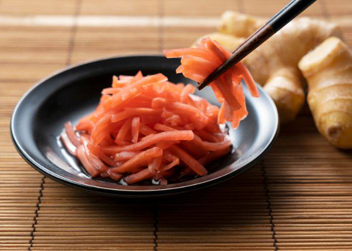 Chopsticks picking up thin slices of ginger. In the background, whole ginger.